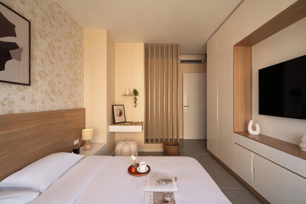 A hotel room with a bed, a bedside table with a lamp, a TV mounted on the wall, and a small shelf with decorative items.