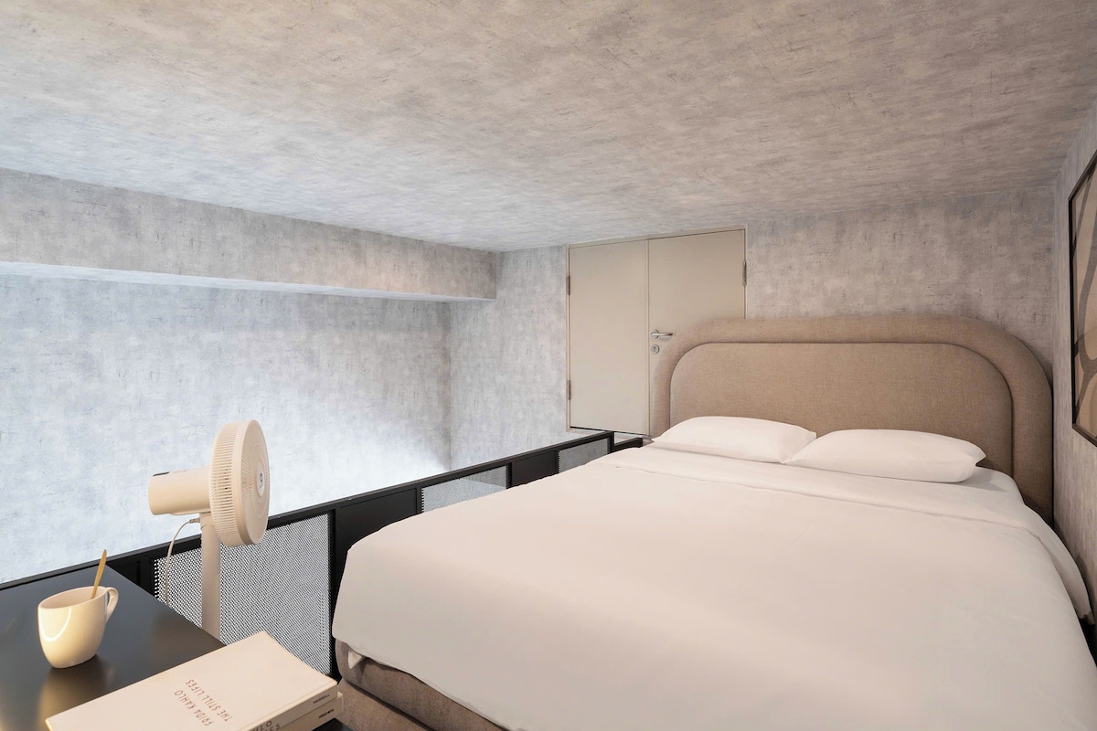 A modern hotel room with a bed, a desk with a lamp, and a bookshelf.