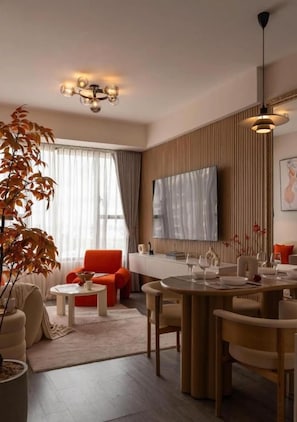 A modern living room with a dining area, a large window with curtains, a flat-screen TV mounted on the wall, and a potted plant. - Rivergate Residence D4 by Veluxe Stay (Khanh Hoi)