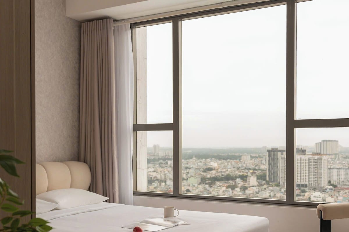 A modern hotel room with a large bed, a view of a cityscape, and a small table with a red apple.