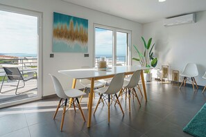 A modern dining area with a white table and chairs, a large painting on the wall, and a view of the sea through the windows. - Silver Villa with private pool and sea view (Atalaia)