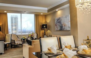 Habitación de hotel con zona de comedor, sofá, mesa lista para una comida y vistas de un paisaje urbano a través de amplias ventanas.