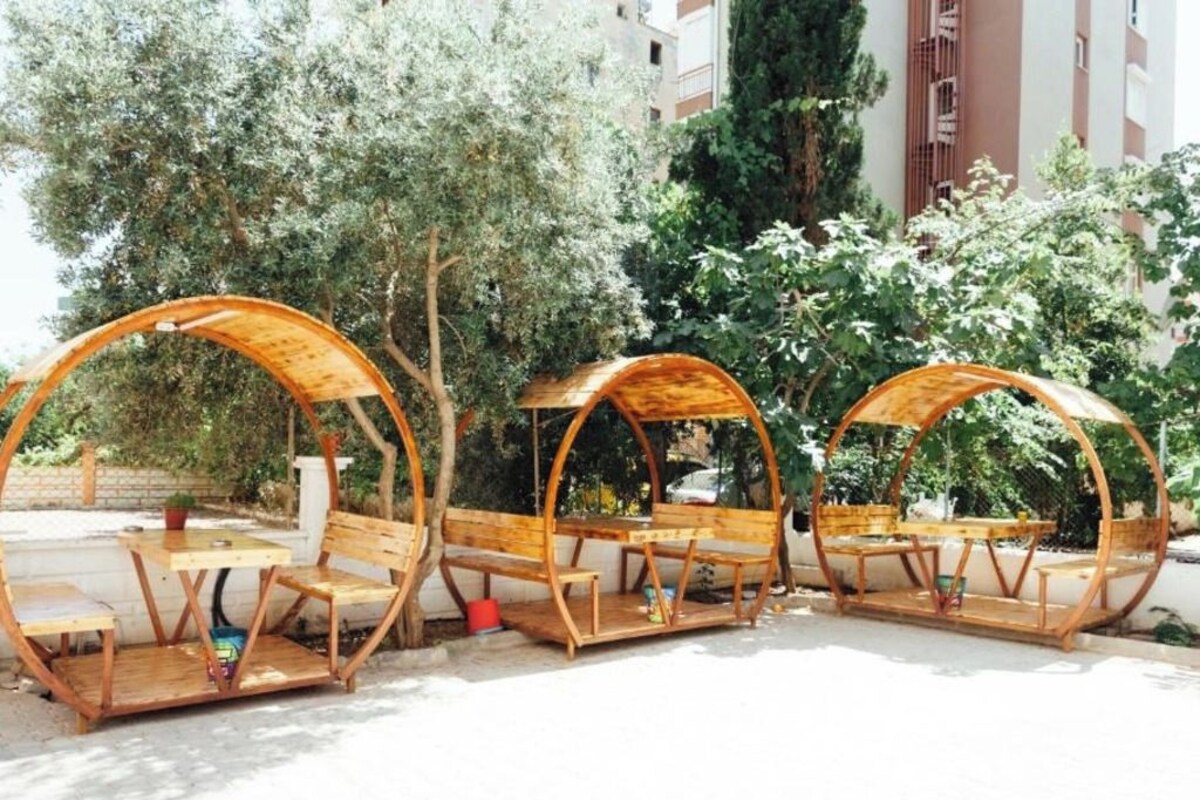 Three wooden outdoor seating areas with benches and tables, surrounded by trees and buildings.