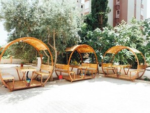 Trois espaces de détente extérieurs en bois, dotés de bancs et de tables, et entourés d’arbres et de bâtiments.