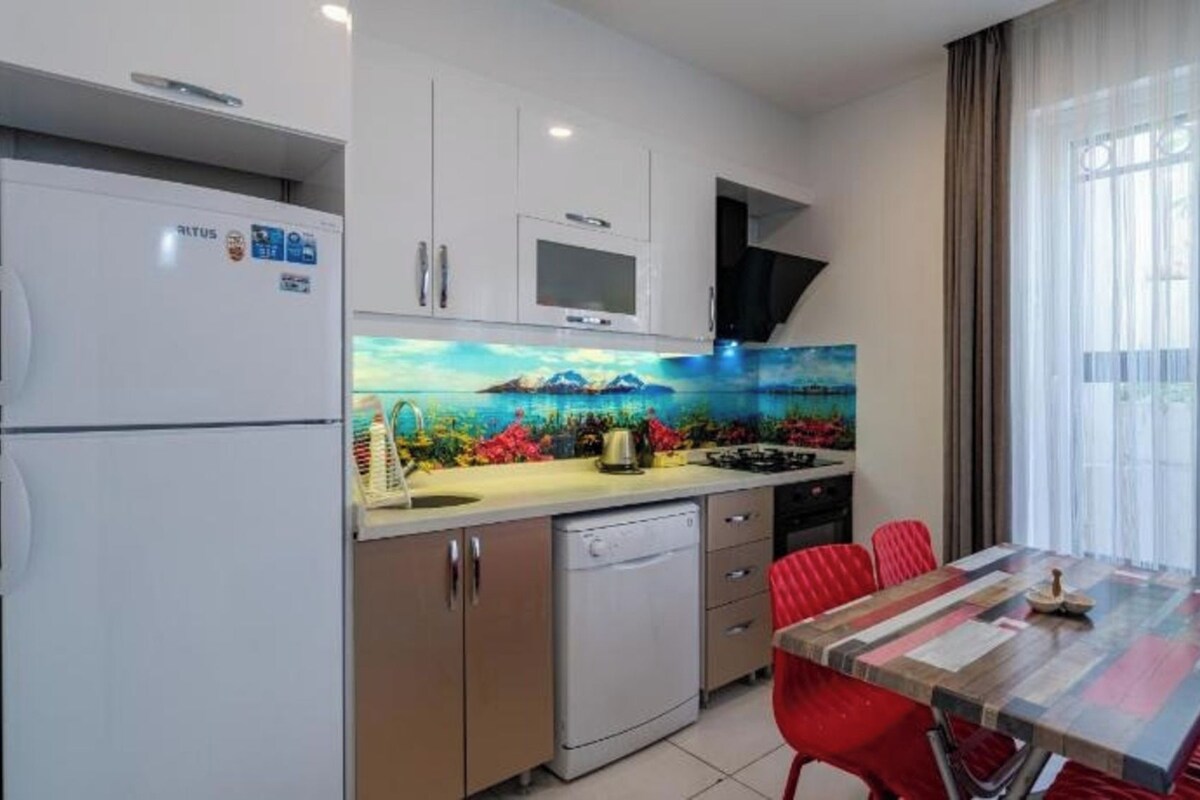 A compact kitchen with a refrigerator, microwave, and a dishwasher. There's a dining table with red chairs and a view through large windows.