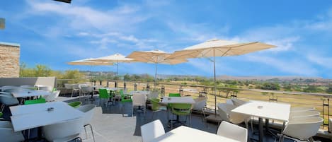 Eine Terrasse im Freien mit Tischen, Stühlen und Sonnenschirmen, mit Blick auf die Landschaft.