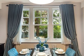 En matplats med ett bord dukat för fyra personer, ett fönster med gardiner och utsikt över grönområden utanför.