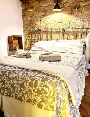 A bed with a patterned bedspread, a stone wall behind it, and a small wooden shelf with a plant. - La Cicala - Charm & Relaxation Just 5 Minutes from the Sea (Rosignano Marittimo)