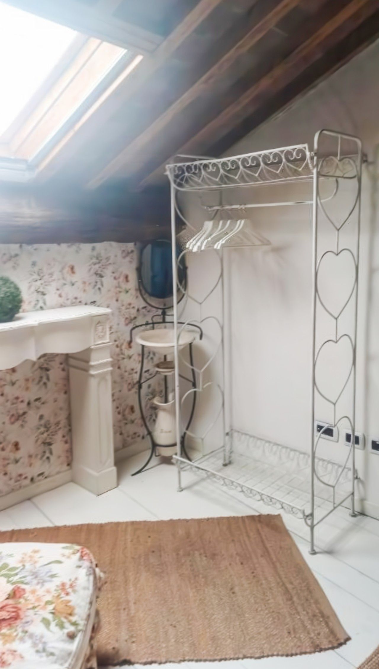 Ein Zimmer mit Dachfenster, einem weißen Metallregal mit Herzformen, einem kleinen weißen Tisch und einem Teppich mit Blumenmuster.