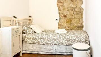 A single bed with a patterned bedspread, a bedside table, a small cabinet, and a stone wall.