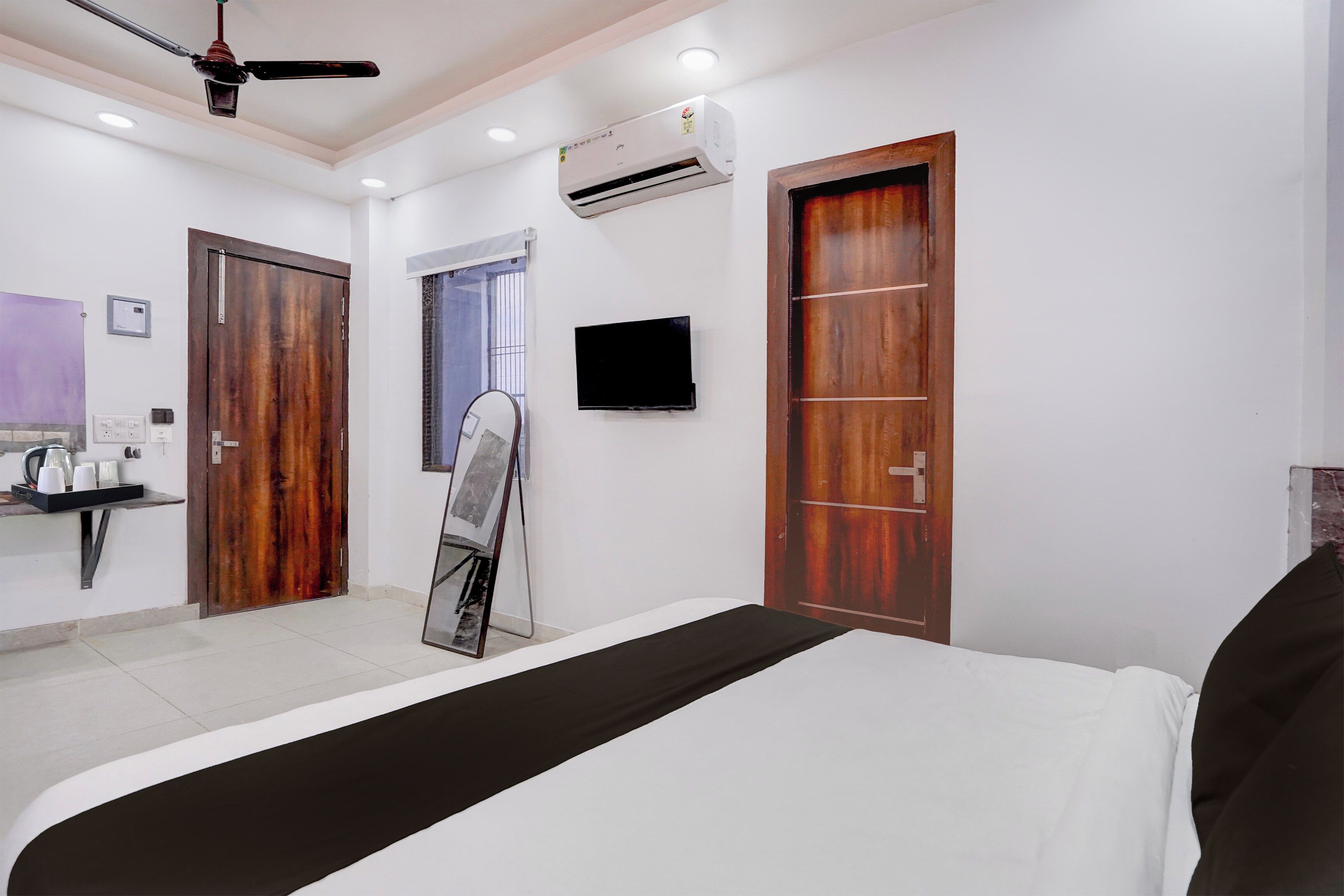Habitación de hotel con cama, ventilador de techo, unidad de aire acondicionado, televisor, espejo y puerta de madera.