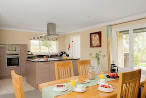 A modern kitchen with a dining area, featuring a central island, built-in appliances, and a view through large windows. - The Allensford (Hexham)