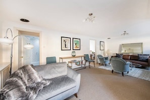 A living room with a grey sofa, a coffee table, and a bookshelf. There are two chairs, a mirror, and a chandelier. - 2 Bed Apartment, Notting Hill (London)