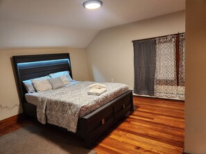 A bedroom with a bed, a headboard, a bedside table, a nightstand, a window with curtains, and a wooden floor. - Blue siren nest (Ludlow)