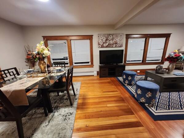 A dining area with a glass-top table, chairs, a vase with flowers, and a television in the background.