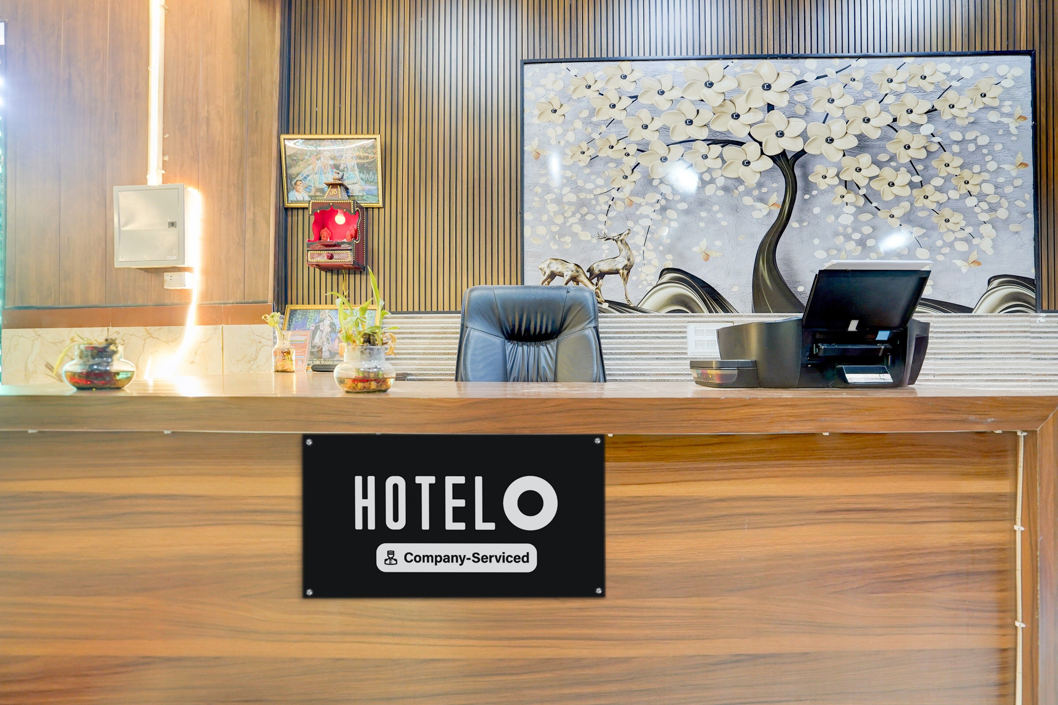 Mostrador de recepción de hotel con un cartel que indica servicio de empresa. Hay una foto en un marco en la pared, un mural decorativo de un árbol y una estatua de ciervo.