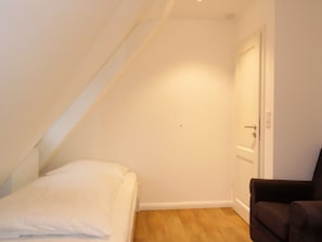 A small, narrow room with a sloped ceiling, a single bed with white bedding, a brown chair, and a closed door. - 4 Caymans at Boldix House (Wyk auf Foehr)