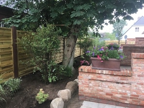 A garden with a wooden fence, a tree, a brick planter with flowers, and a brick wall. - 4 Caymans at Boldix House (Wyk auf Foehr)