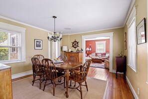A dining area with a wooden table and chairs, a chandelier, and a view of the outside through large windows. - Historic Ptown Charm - 3BR, Yard, AC, Parking (Provincetown)