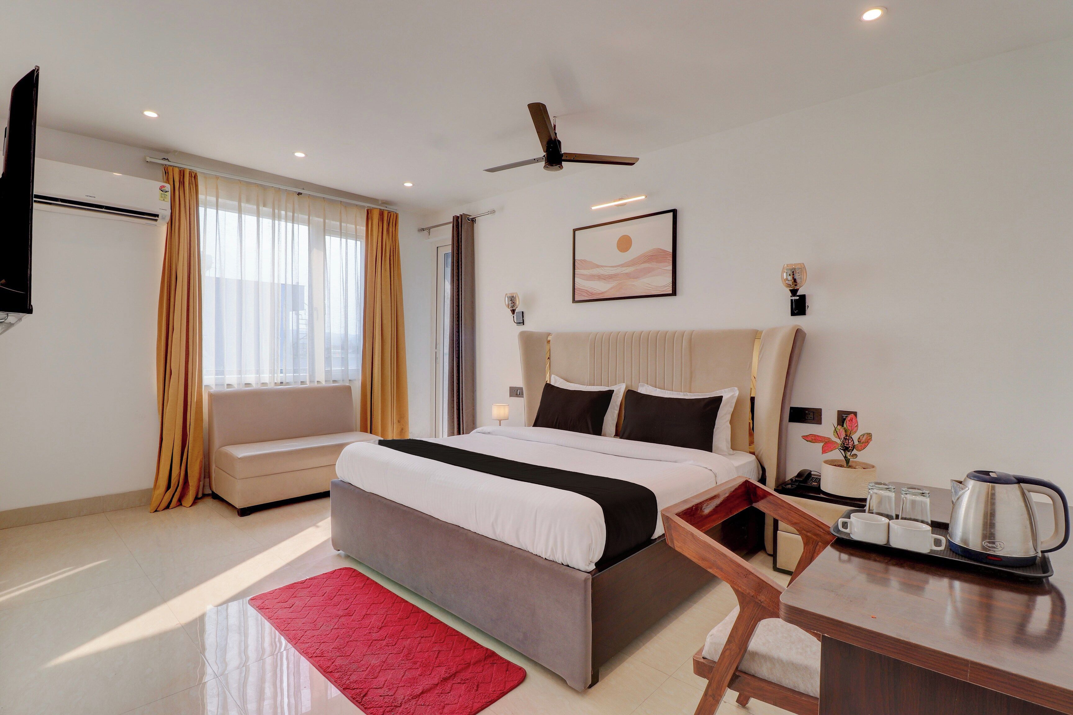 Une chambre d’hôtel avec un lit, un bureau, une chaise, un tapis rouge, un ventilateur de plafond et un tableau au mur.