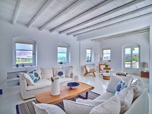 A spacious living room with a high ceiling, white walls, and multiple windows offering a view of the landscape. - Villa Kyma - Greek Summer (Kamares Naousas)
