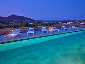 A swimming pool with lounge chairs and a stone wall, overlooking a coastal landscape with buildings and a mountain. - Villa Kyma - Greek Summer (Kamares Naousas)