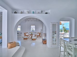 A spacious room with white walls, a central seating area with chairs and a table, a window with a view, and a balcony with outdoor seating. - Villa Kyma - Greek Summer (Kamares Naousas)