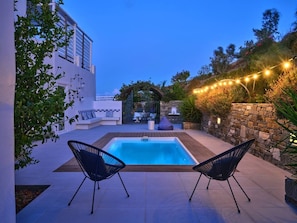A swimming pool with two chairs, surrounded by a stone wall and plants, with string lights in the background. - Villa Kyma - Greek Summer (Kamares Naousas)