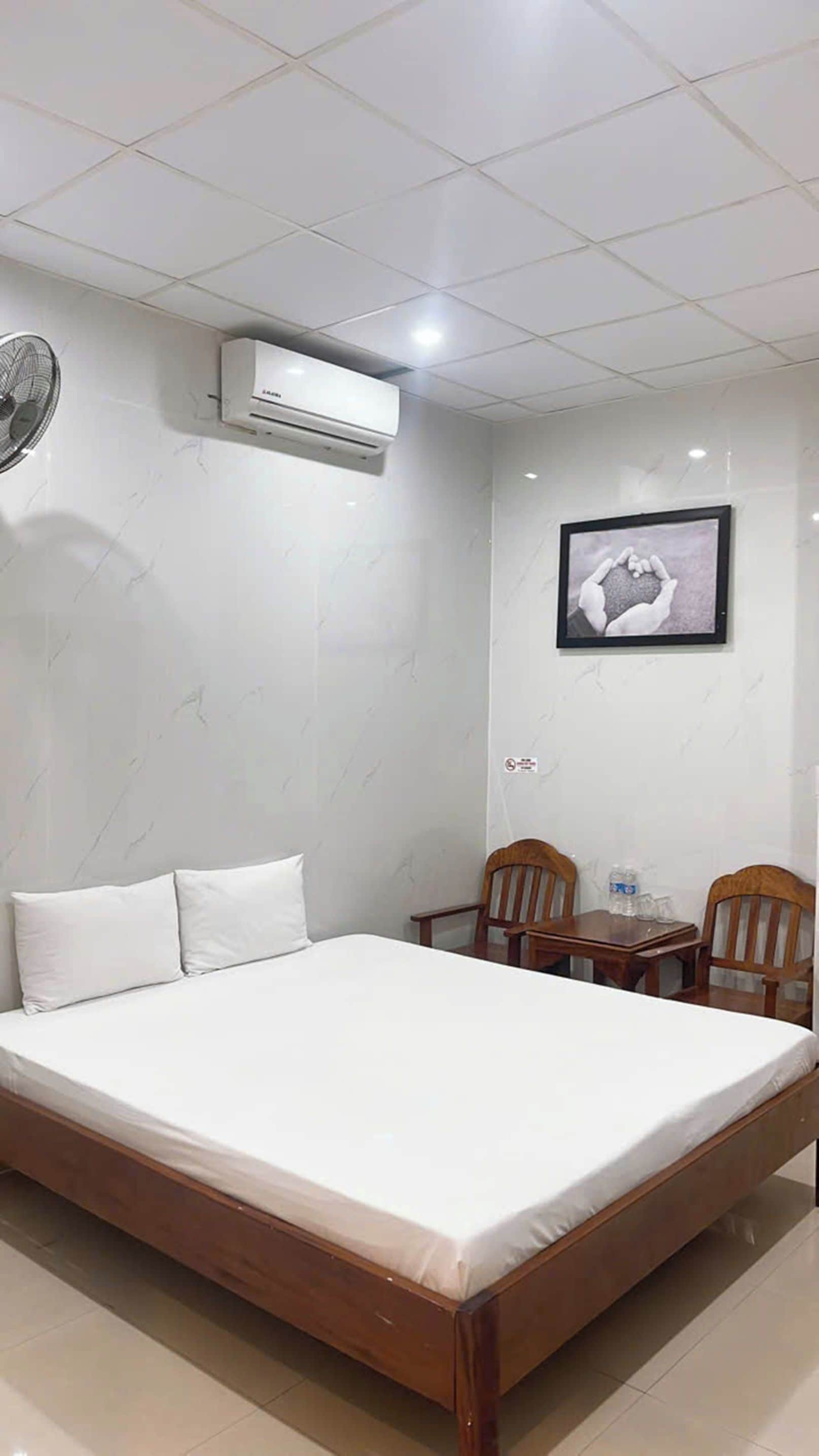 Une chambre d’hôtel avec un lit, deux chaises en bois, une petite table, un appareil de climatisation et un tableau encadré au mur.