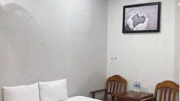 Une chambre d’hôtel avec un lit, deux chaises en bois, une petite table, un appareil de climatisation et un tableau encadré au mur.