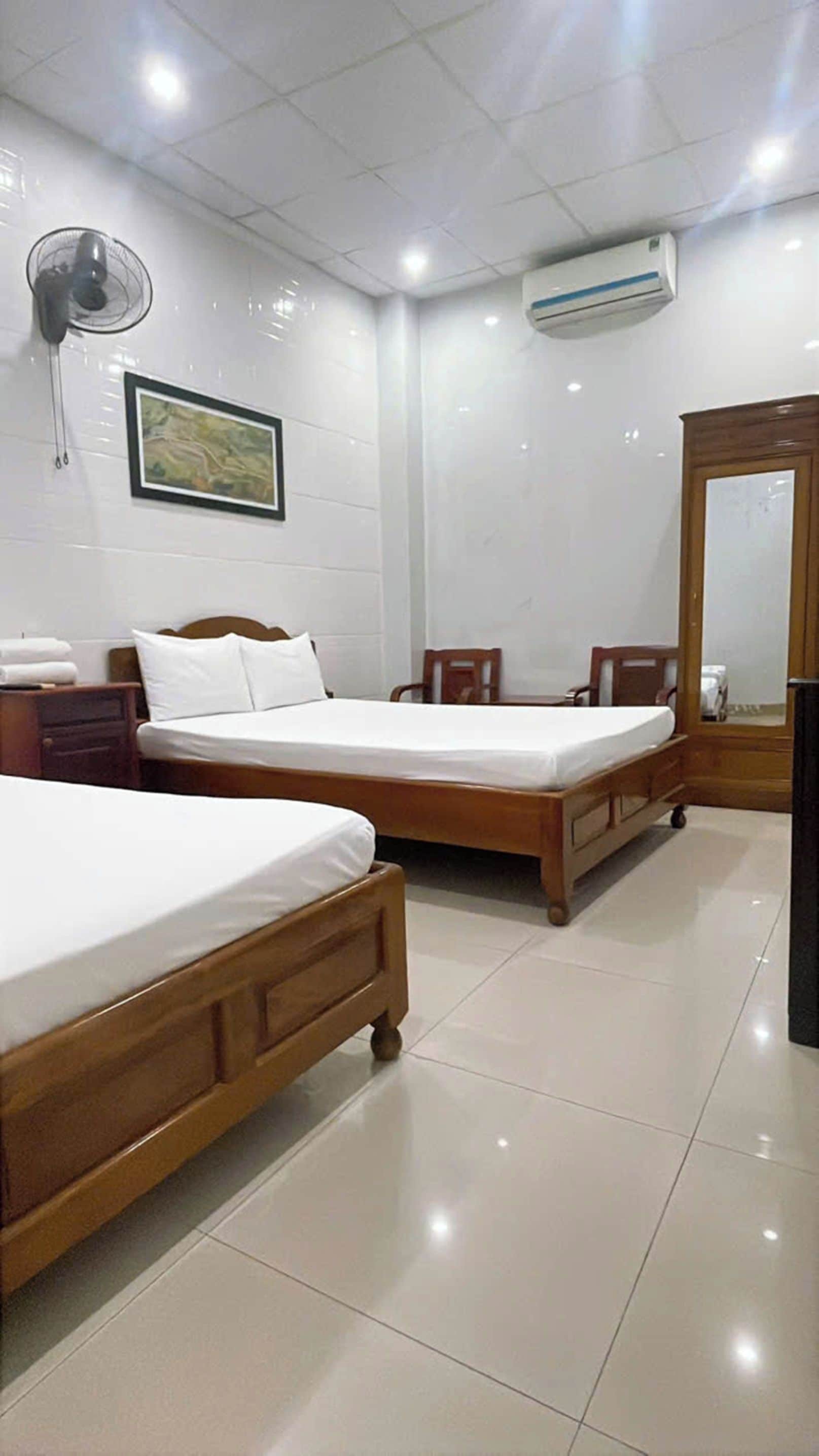 A hotel room with two wooden beds, a bedside table, a chair, a standing fan, an air conditioning unit, and a framed picture on the wall.