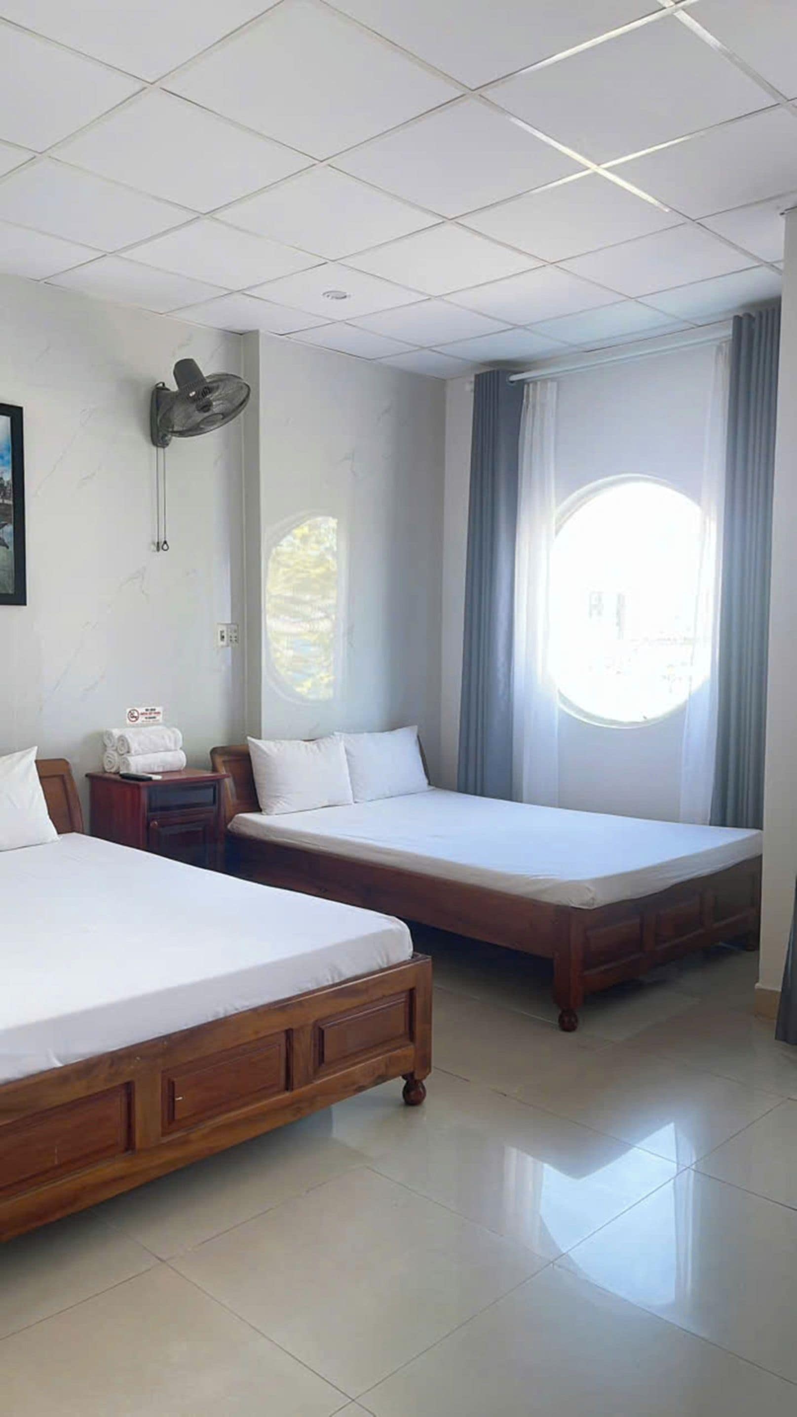 Una camera d'albergo con due letti singoli, una ventola a parete, un comodino con un telecomando e una finestra con tende.
