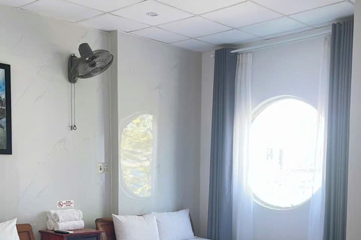 A hotel room with two single beds, a wall-mounted fan, a bedside table with a remote control, and a window with curtains.