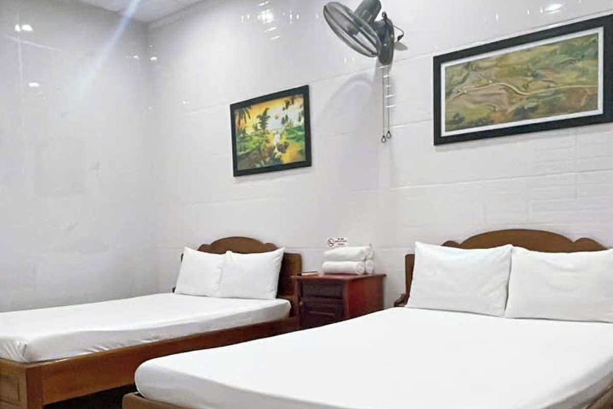 A hotel room with two wooden beds, white bedding, a wall-mounted fan, and two framed pictures.