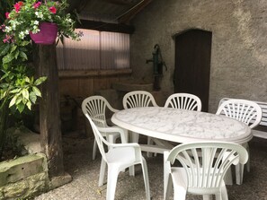 An outdoor patio with a round marble table and white plastic chairs, surrounded by greenery and a rustic stone wall. - New Listing (Desseling)