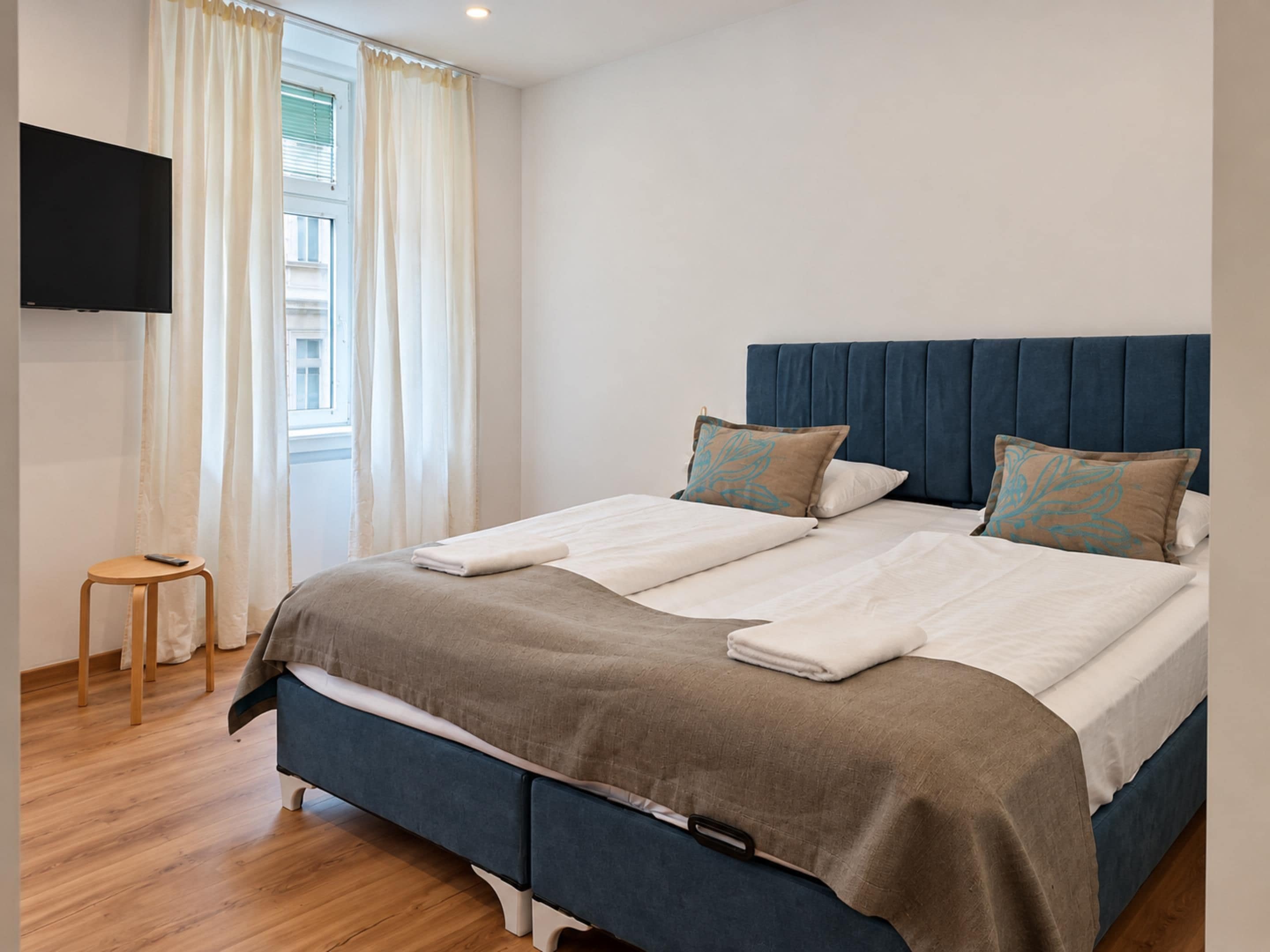 A neatly made bed with a blue headboard, a remote control on a wooden stool, and a television mounted on the wall.