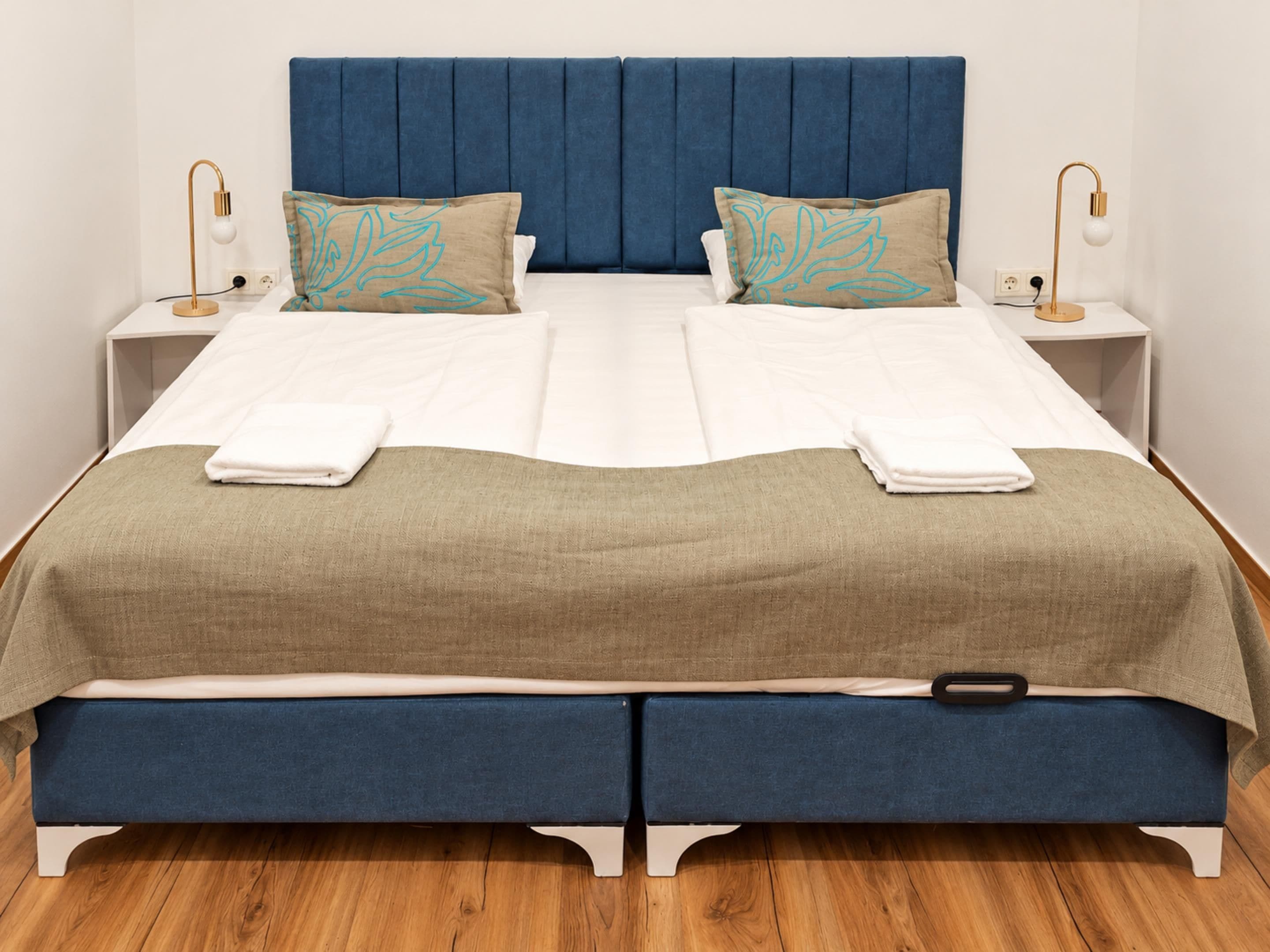 Una cama doble con cabecera azul, dos mesitas de noche con lámparas y una colcha azul y beige.