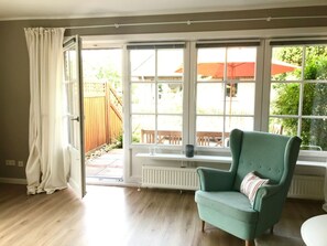 A room with a teal armchair, a window with a view of a wooden deck and outdoor furniture, and a radiator under the window. - View of the Dike in Nieblum (Nieblum)