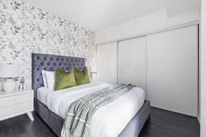 A bedroom with a tufted headboard, a bed with white bedding, a grey patterned throw, and a nightstand with a lamp. - Charming 2BR downtown condo with private patio Parking Available (Toronto)