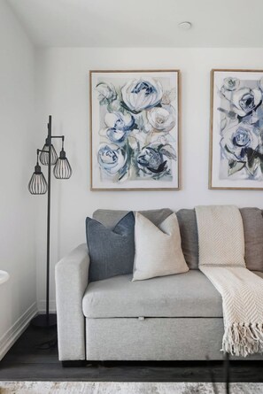A living room with a grey sofa, multiple cushions, and two framed floral paintings on the wall. - Charming 2BR downtown condo with private patio Parking Available (Toronto)