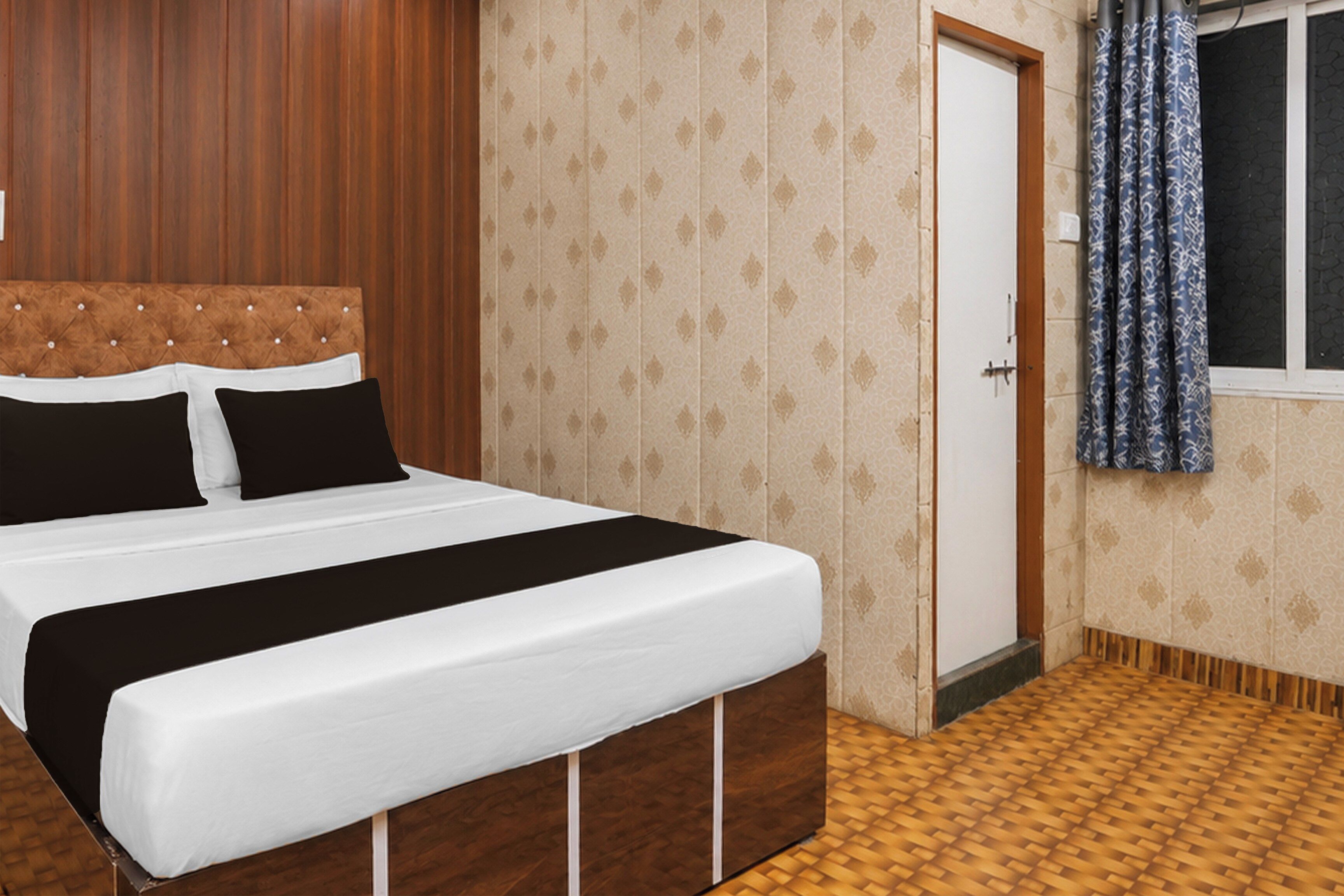 Una habitación de hotel con una cama, cabecera de madera, papel tapiz estampado, una puerta y una ventana con cortinas.