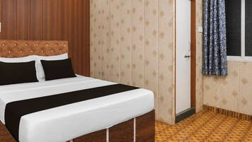 Una habitación de hotel con una cama, cabecera de madera, papel tapiz estampado, una puerta y una ventana con cortinas.