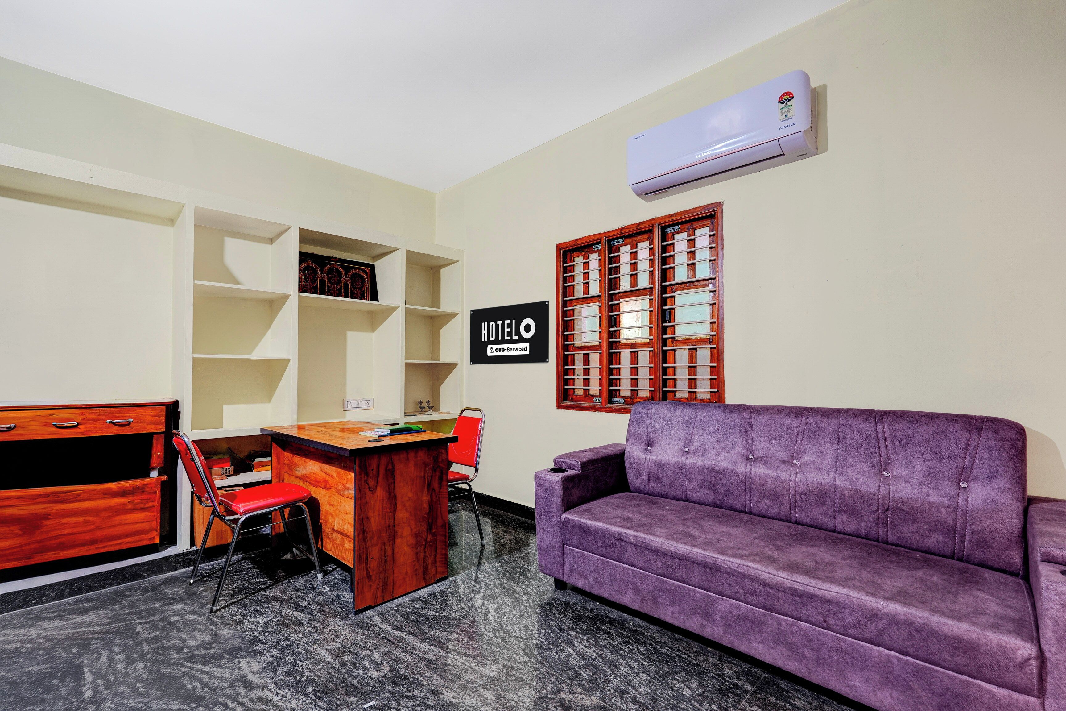 Une chambre d’hôtel avec un bureau en bois, un canapé violet, une chaise rouge, une bibliothèque et un panneau indiquant que la chambre est desservie par le service de l’hôtel.