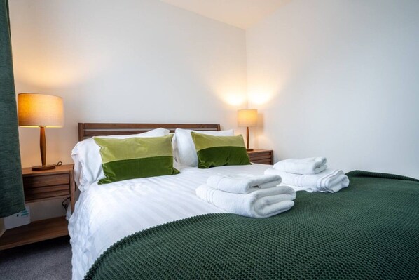 A neatly made bed with white bedding and green pillows, two bedside lamps, and a green throw blanket.
