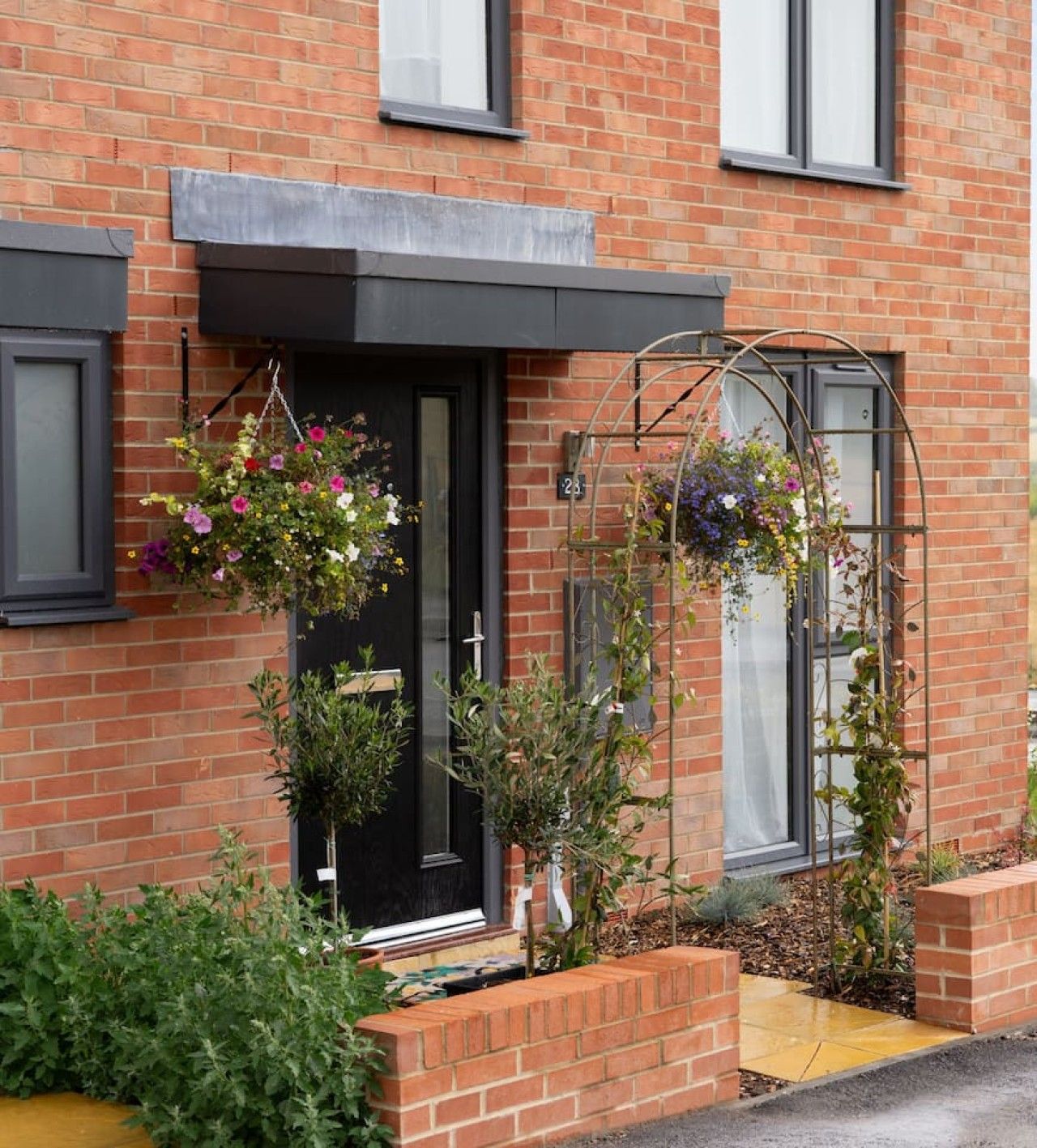 Une maison en briques avec une porte noire, un petit jardin agrémenté de plantes en pot et un treillage métallique orné de fleurs suspendues.