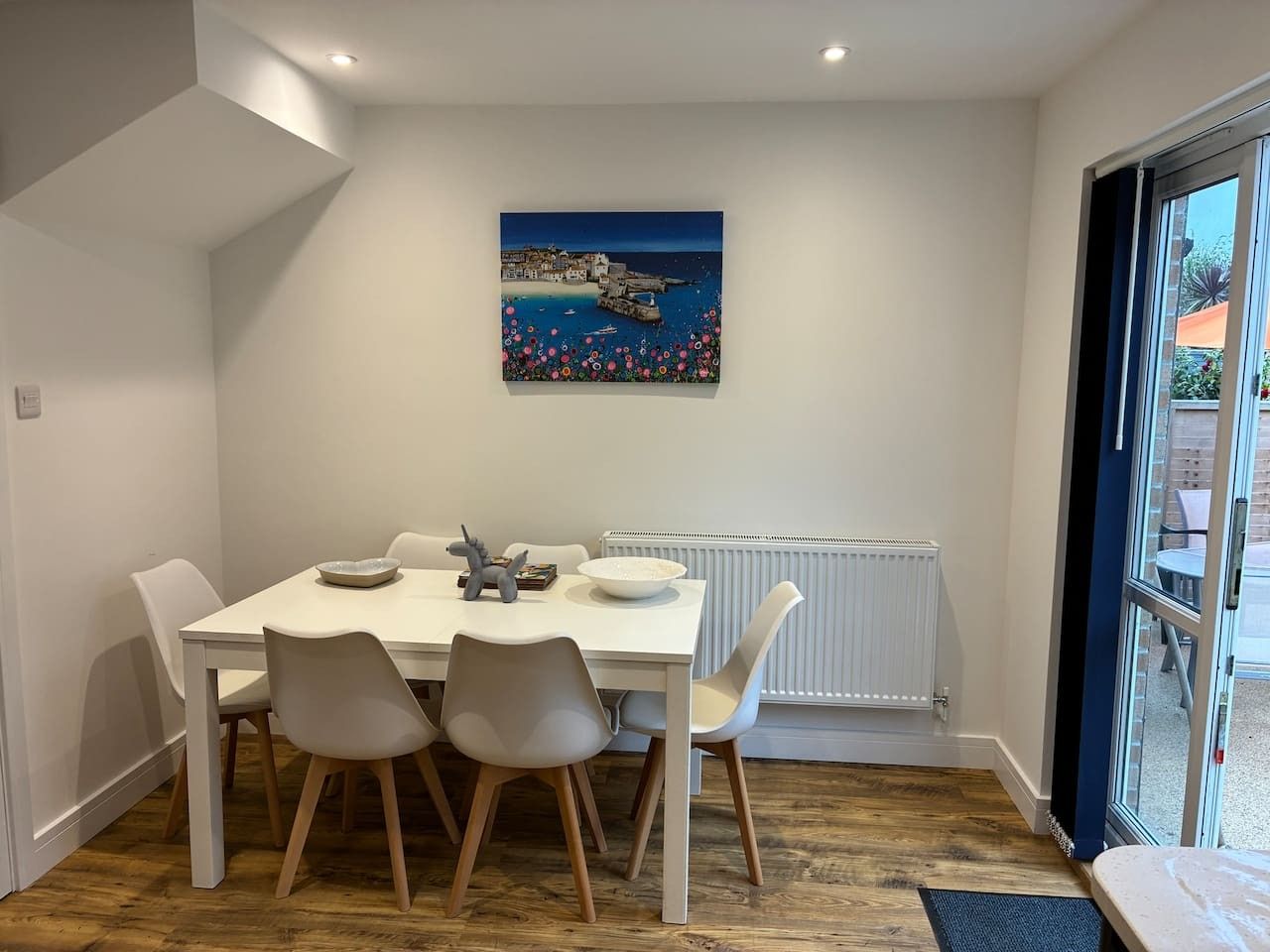 A dining area with a white table and chairs, a painting on the wall, and a view through a glass door.