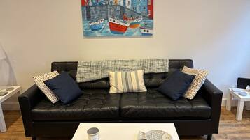 Ein schwarzes Ledersofa mit gemusterten Kissen, ein weißer Couchtisch mit einem fischförmigen Gegenstand und ein farbenfrohes Gemälde von Booten an der Wand.