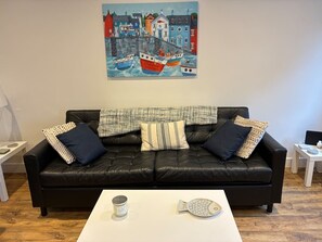 Un canapé en cuir noir agrémenté de coussins à motifs, une table basse blanche sur laquelle repose un objet en forme de poisson, et un tableau coloré représentant des bateaux accroché au mur.