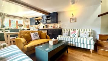 A cozy living room with a sofa, a striped armchair, and a coffee table. There is a kitchen area with cabinets and a window.