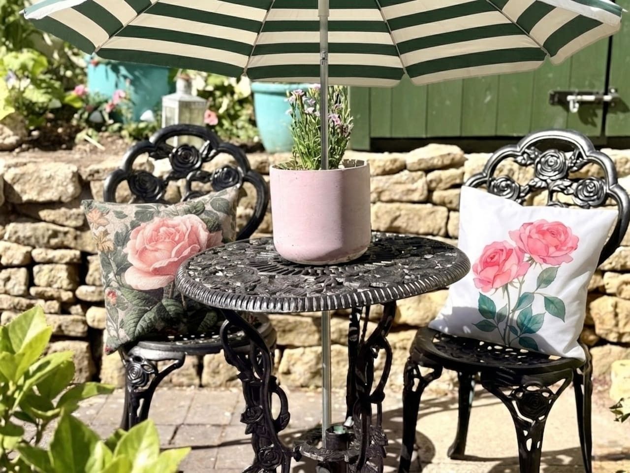 A round metal table with a potted plant, two chairs with floral cushions, and a striped umbrella.
